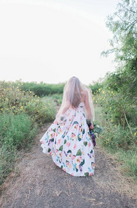 A woman viewed from behind walking on a dirt path outdoors, wearing a flowing white dress with a colorful floral print and holding a bouquet of purple flowers.