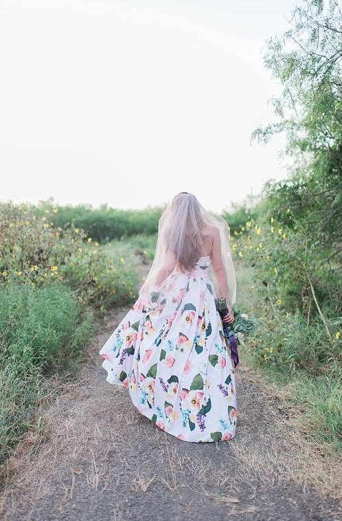 A woman viewed from behind walking on a dirt path outdoors, wearing a flowing white dress with a colorful floral print and holding a bouquet of purple flowers.