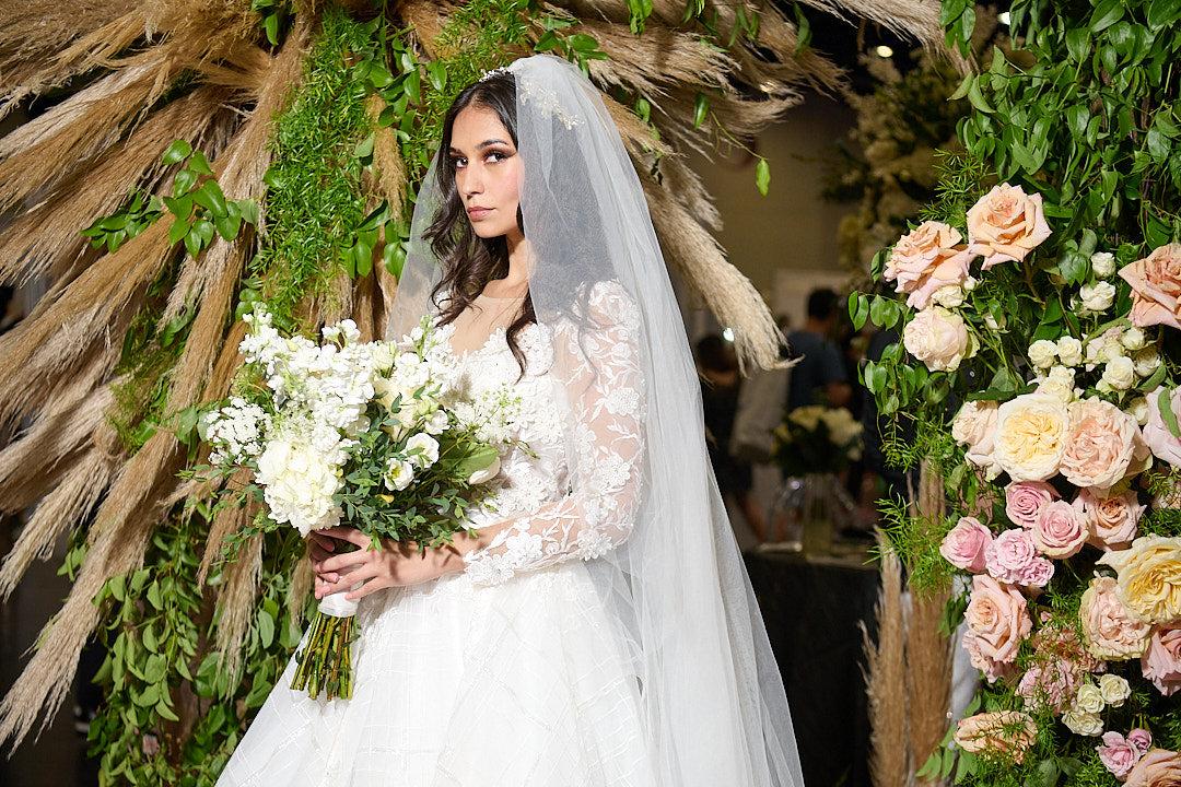 Bride in custom lace wedding dress with veil, holding white bouquet, surrounded by greenery and pink roses