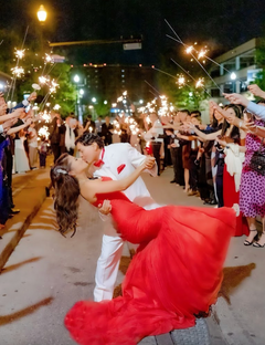 Bride in bold red wedding dress kissed by groom under sparklers at night, celebrating love with guests