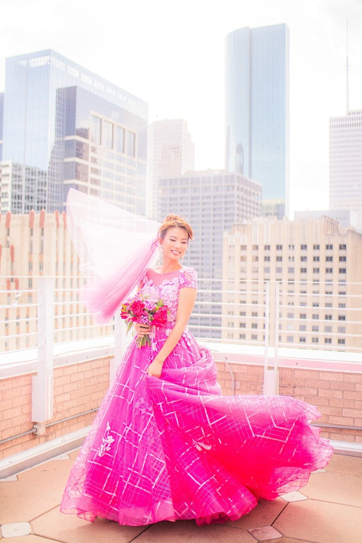 Bride in vibrant pink custom wedding dress with city skyline, holding bouquet, veil flowing