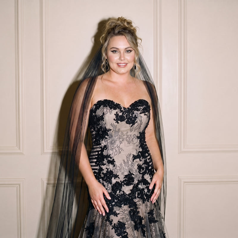 A woman wearing a black lace strapless gown with a sheer black tulle overlay and veil, standing against a beige paneled wall.