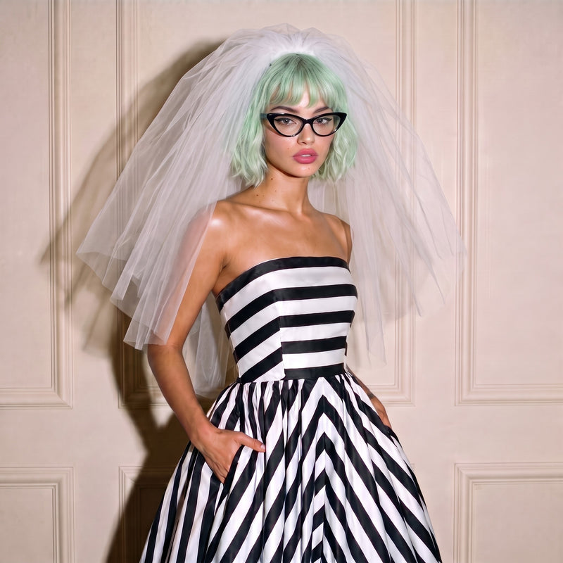 A model wearing a black and white striped strapless wedding dress a white tulle bridal veil, and mint green hair, posing in front of a beige paneled wall.
