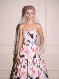 A woman wearing a strapless pink floral ball gown wedding dress with a colorful floral print and a sheer veil, standing against an off-white paneled wall.