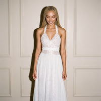 A woman wearing a white lace halter wedding dress with a chiffon overlay skirt and floral print, standing against a cream-colored paneled wall.