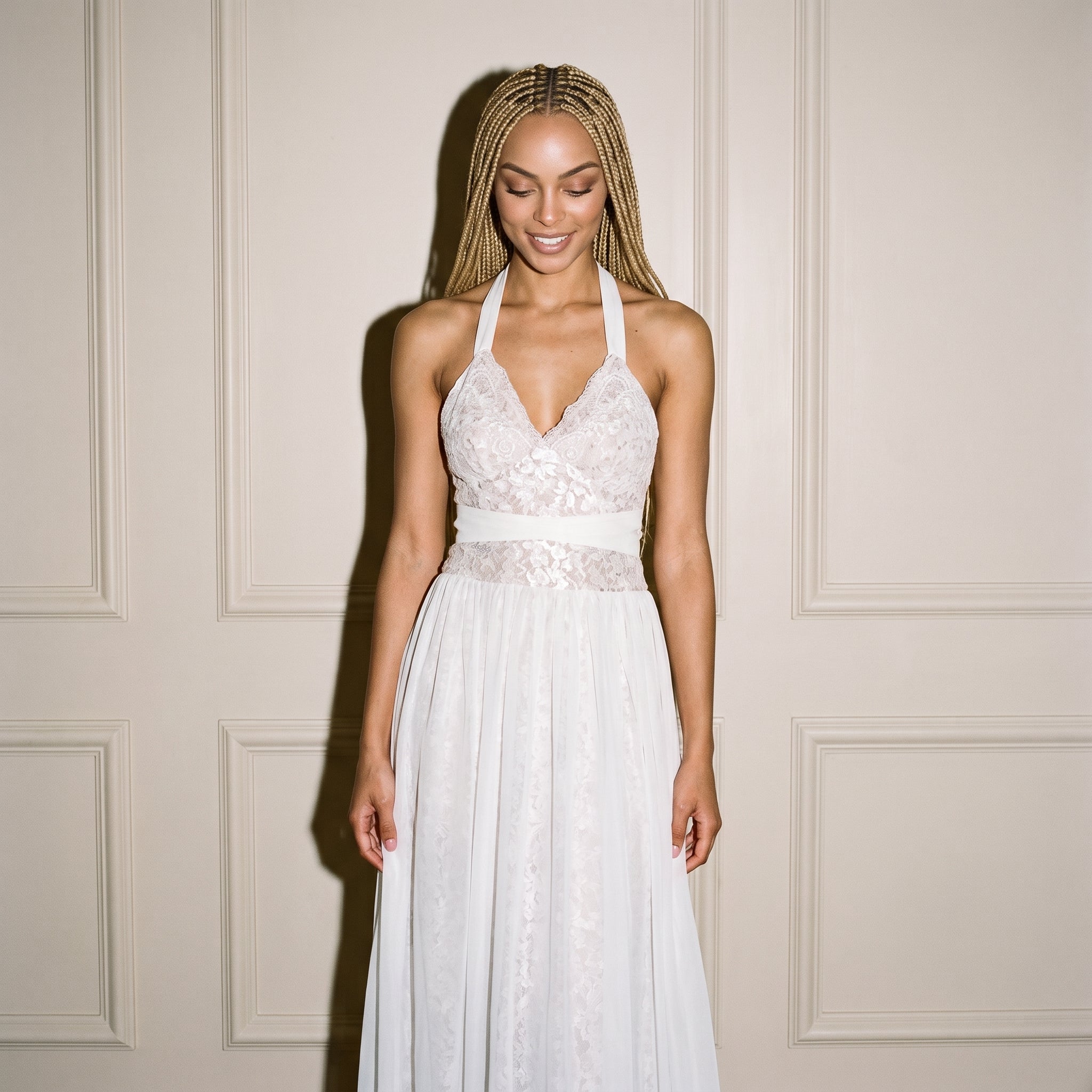 A woman wearing a white lace halter wedding dress with a chiffon overlay skirt and floral print, standing against a cream-colored paneled wall.
