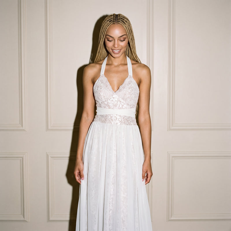 A woman wearing a white lace halter wedding dress with a chiffon overlay skirt and floral print, standing against a cream-colored paneled wall.