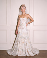 A model wearing a floral print strapless sweetheart mermaid wedding gown with a sheer veil, standing against a white paneled wall.