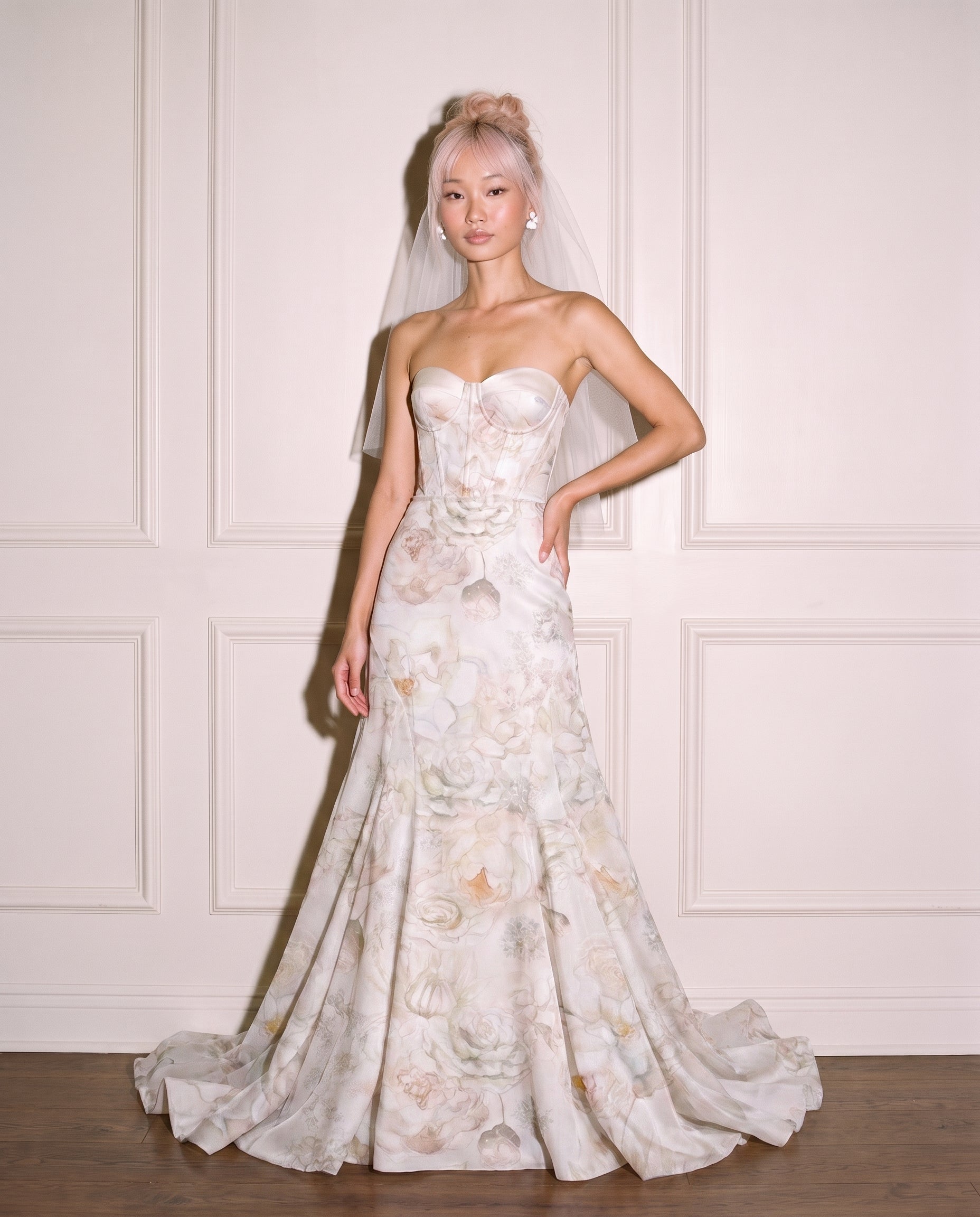 A model wearing a floral print strapless sweetheart mermaid wedding gown with a sheer veil, standing against a white paneled wall.
