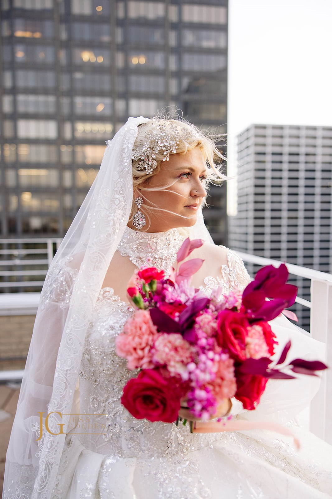 Bride in custom beaded wedding gown with veil, holding vibrant pink and red bouquet on city rooftop