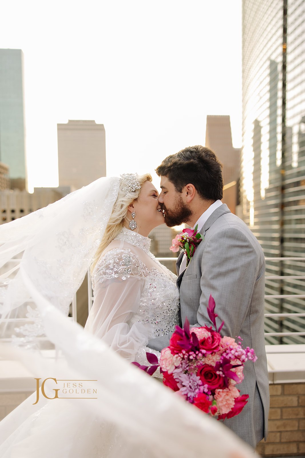 Bride in beaded wedding gown with flowing veil shares tender kiss with groom against city skyline, holding vibrant bouquet