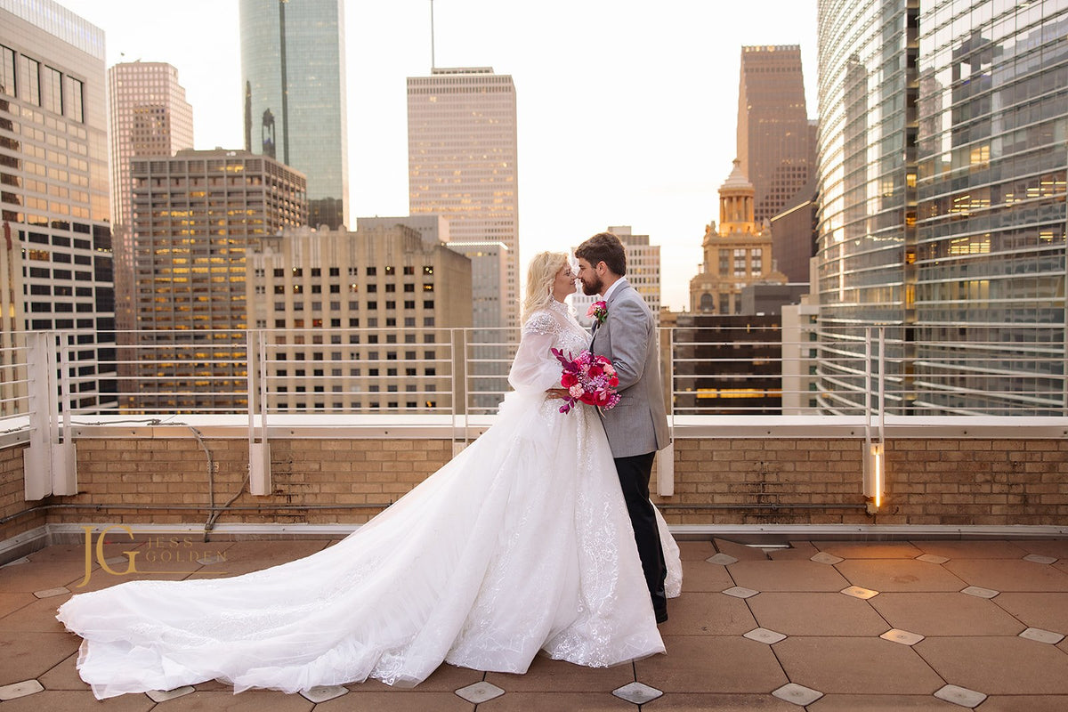 Bride in custom lace wedding gown with long train, embracing groom on rooftop with city skyline at sunset