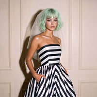A model with mint green hair and a white veil wears a strapless black and white striped wedding dress with a flared skirt, standing against a beige paneled wall.