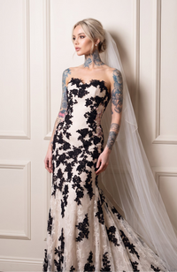 A woman modeling a strapless sweetheart mermaid-style gown with black floral lace applique over a warm ivory base, wearing a long sheer veil. She is posed against an off-white paneled wall.