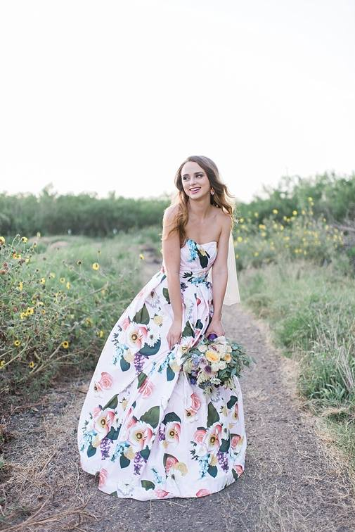 A woman stands outdoors on a dirt path wearing a strapless white bridal gown with a colorful floral print and holding a bouquet of flowers.