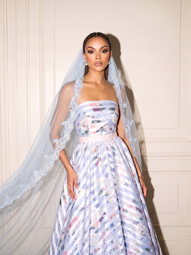 A woman modeling a strapless wedding dress with a blue pastel lace veil. The dress features a pastel blue and white striped pattern with multicolored floral accents and a light pink waistband, set against a cream paneled wall.