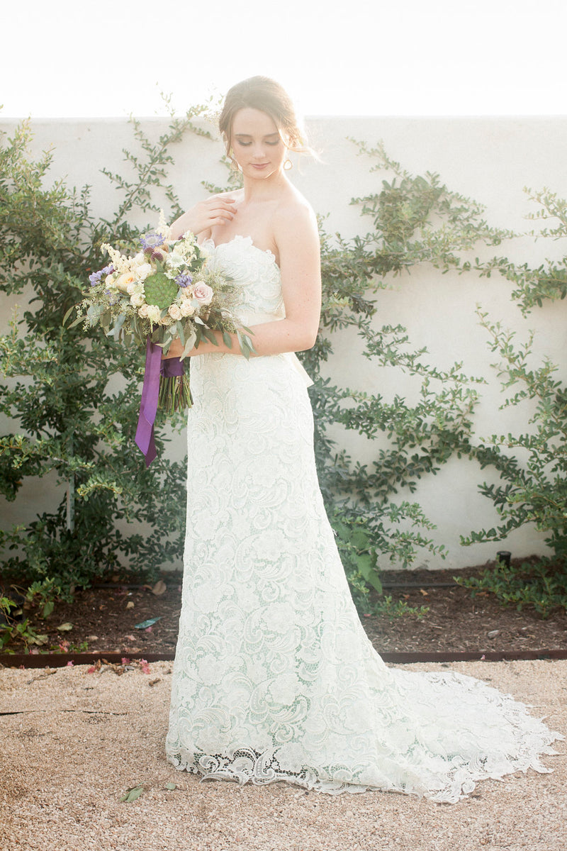 Bride in lace wedding dress holding floral bouquet with purple ribbon, garden setting