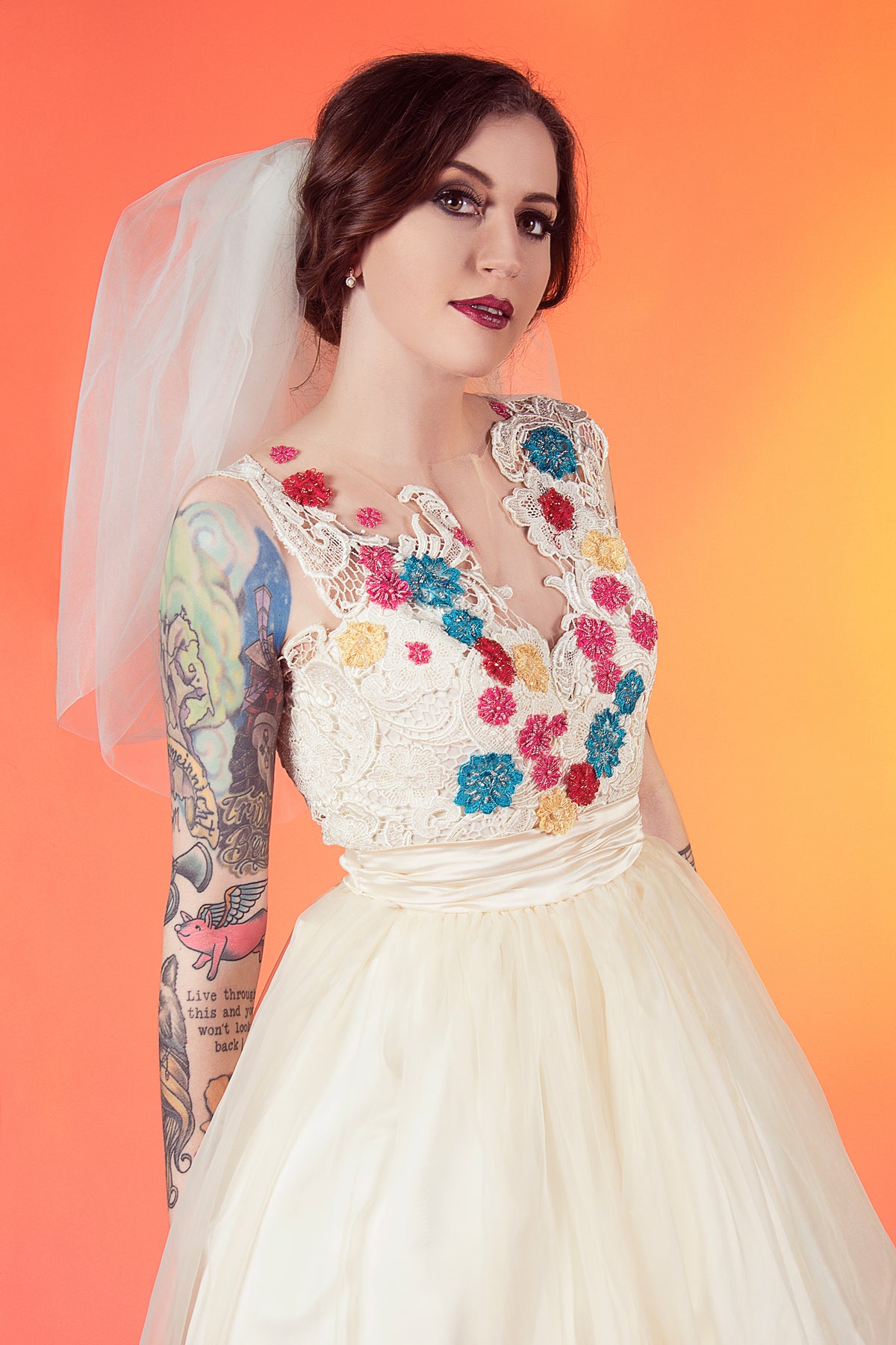 Bride in custom lace wedding dress with colorful floral embroidery, veil, and tattooed arm against orange background