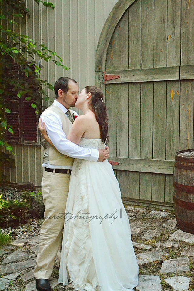 Bride and groom sharing a kiss in front of rustic wooden barn door, wearing custom wedding attire.