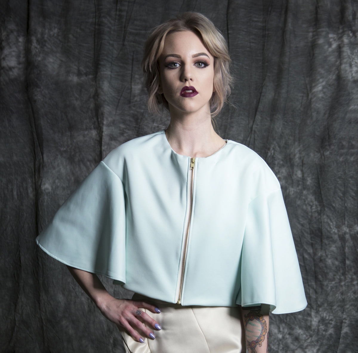 Mint green cape-style top with gold zipper, paired with cream pants, for modern bridal or event wear.