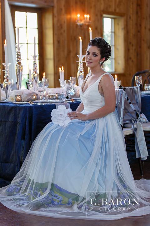 Bride in custom ombre wedding dress with blue-to-white gradient, holding white flower at rustic reception table