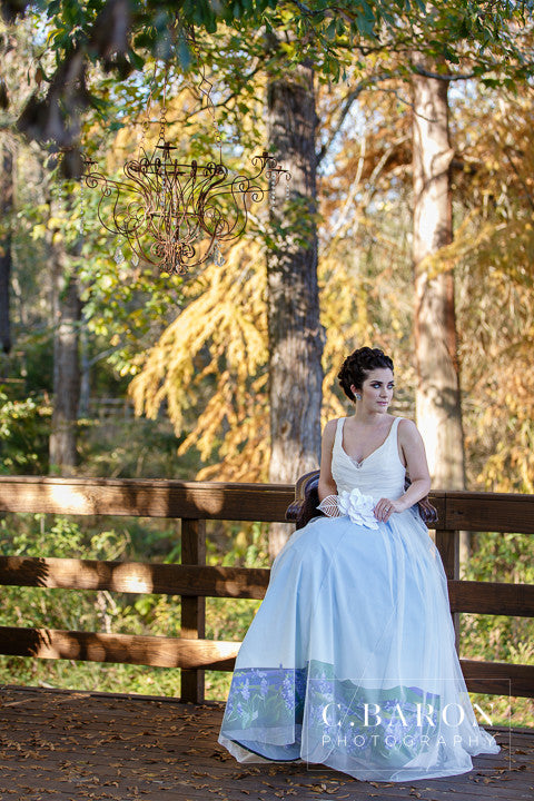 Bride in custom ombre wedding dress with floral hem, seated on wooden bridge under chandelier in autumn forest