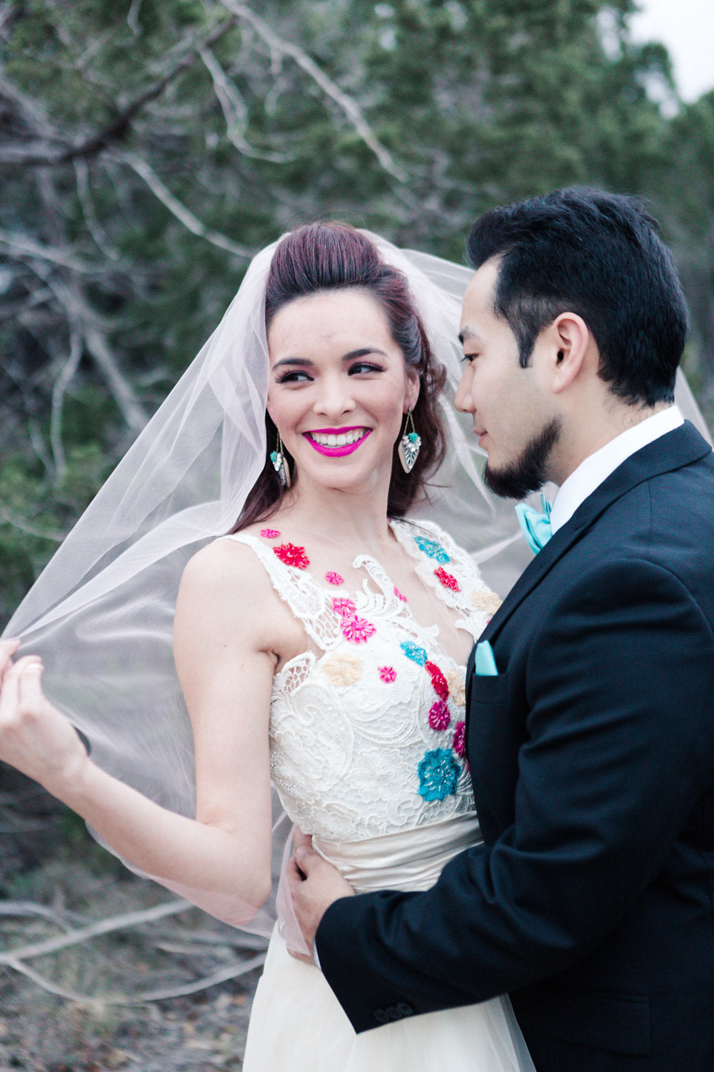 Bride in colorful floral lace wedding dress with veil, groom in black suit, outdoor forest setting