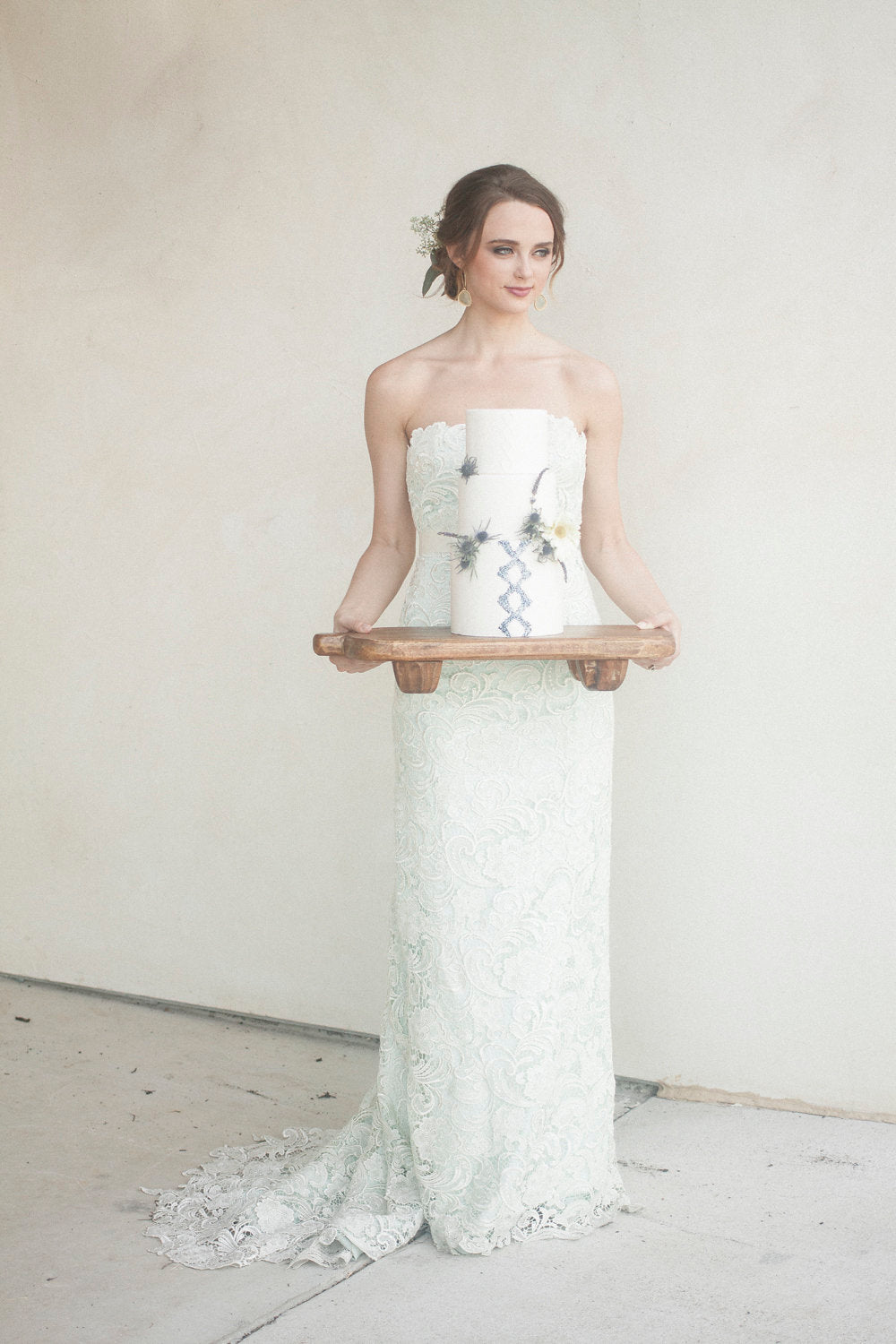 Bride in lace strapless wedding dress holding floral cake on wooden tray