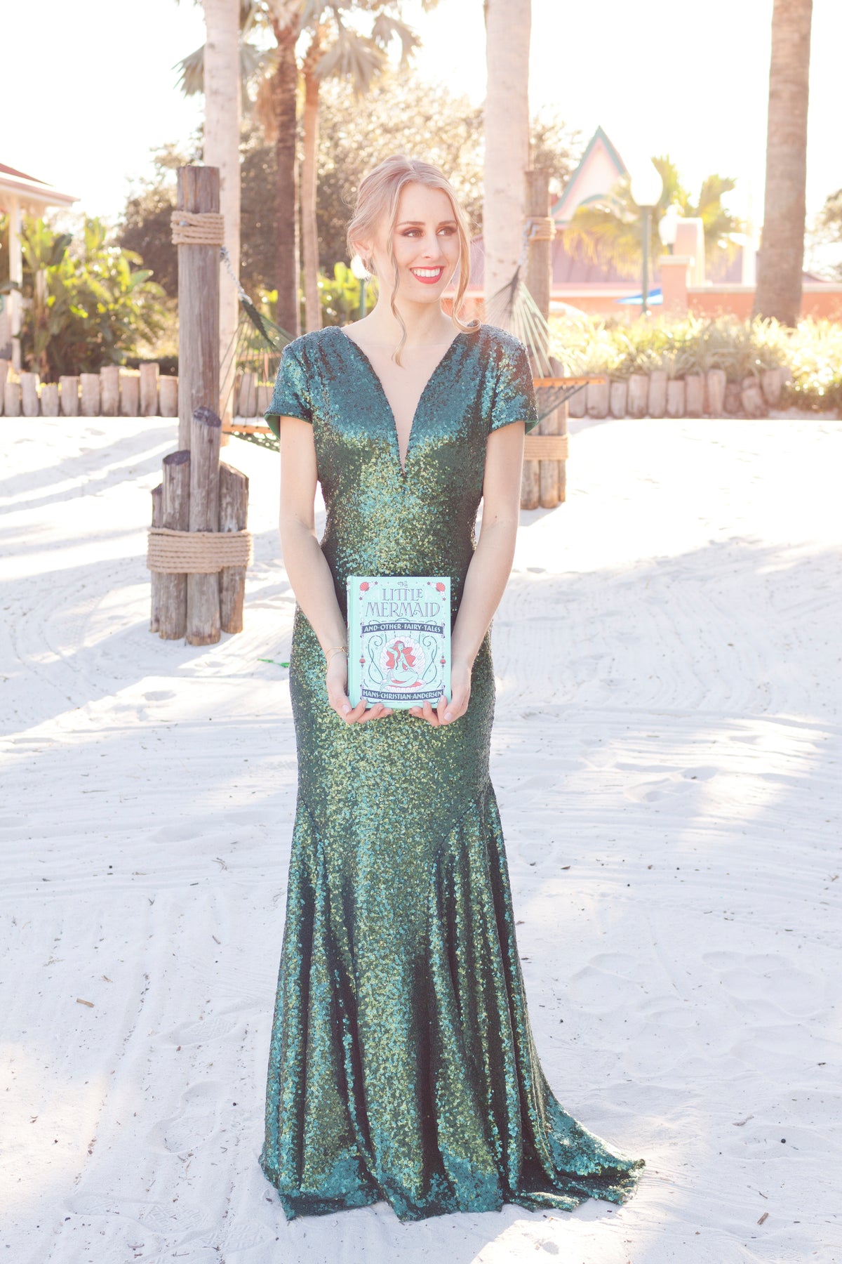 Woman in teal sequin mermaid gown holding 'The Little Mermaid' book on sandy beach