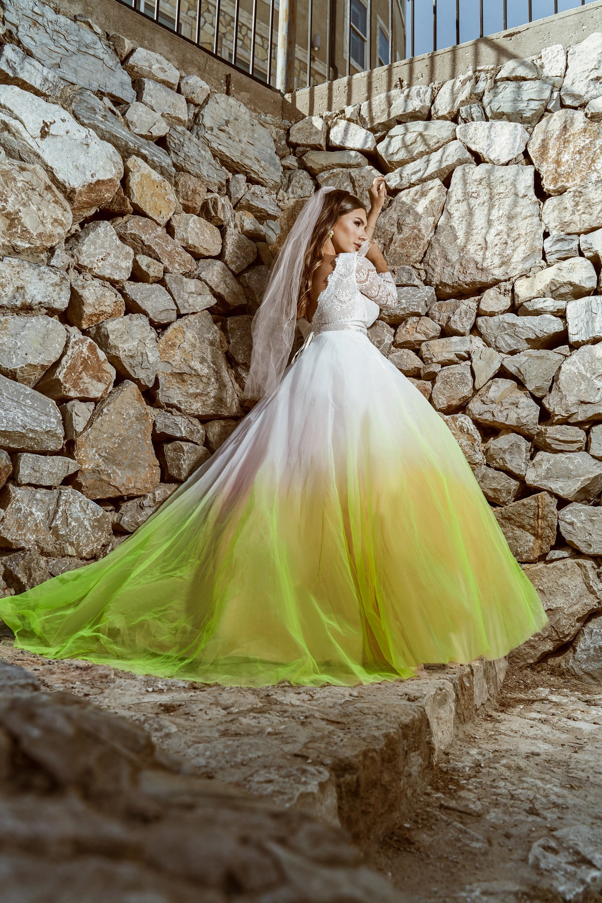Bride in custom ombre wedding dress with neon green skirt, lace sleeves, and veil against stone wall