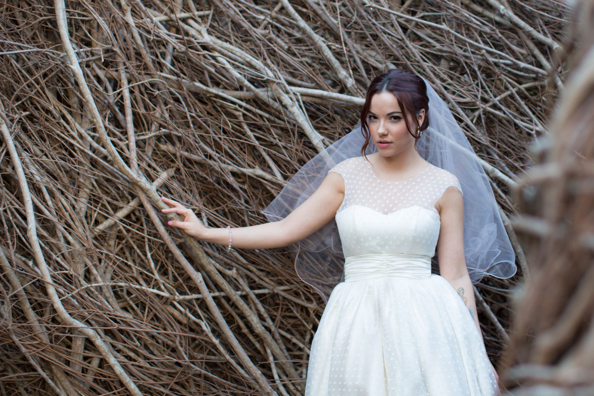 Bride in vintage polka dot wedding dress with veil, standing by rustic twig wall
