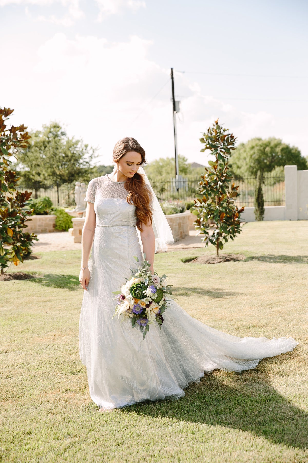 Bride in custom wedding dress with floral bouquet, standing on grassy lawn