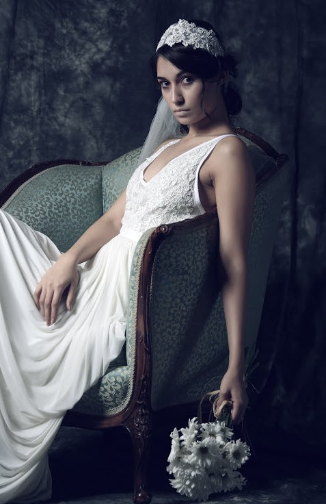 Bride in white lace wedding dress with floral crown, seated on vintage chair holding bouquet