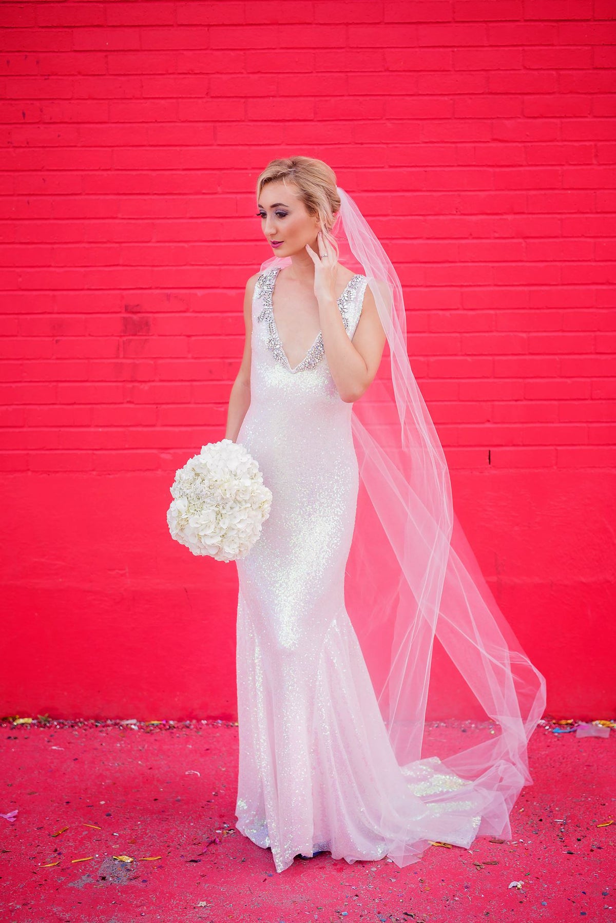 Sparkling white wedding dress with beaded neckline, veil, and bouquet against red brick wall