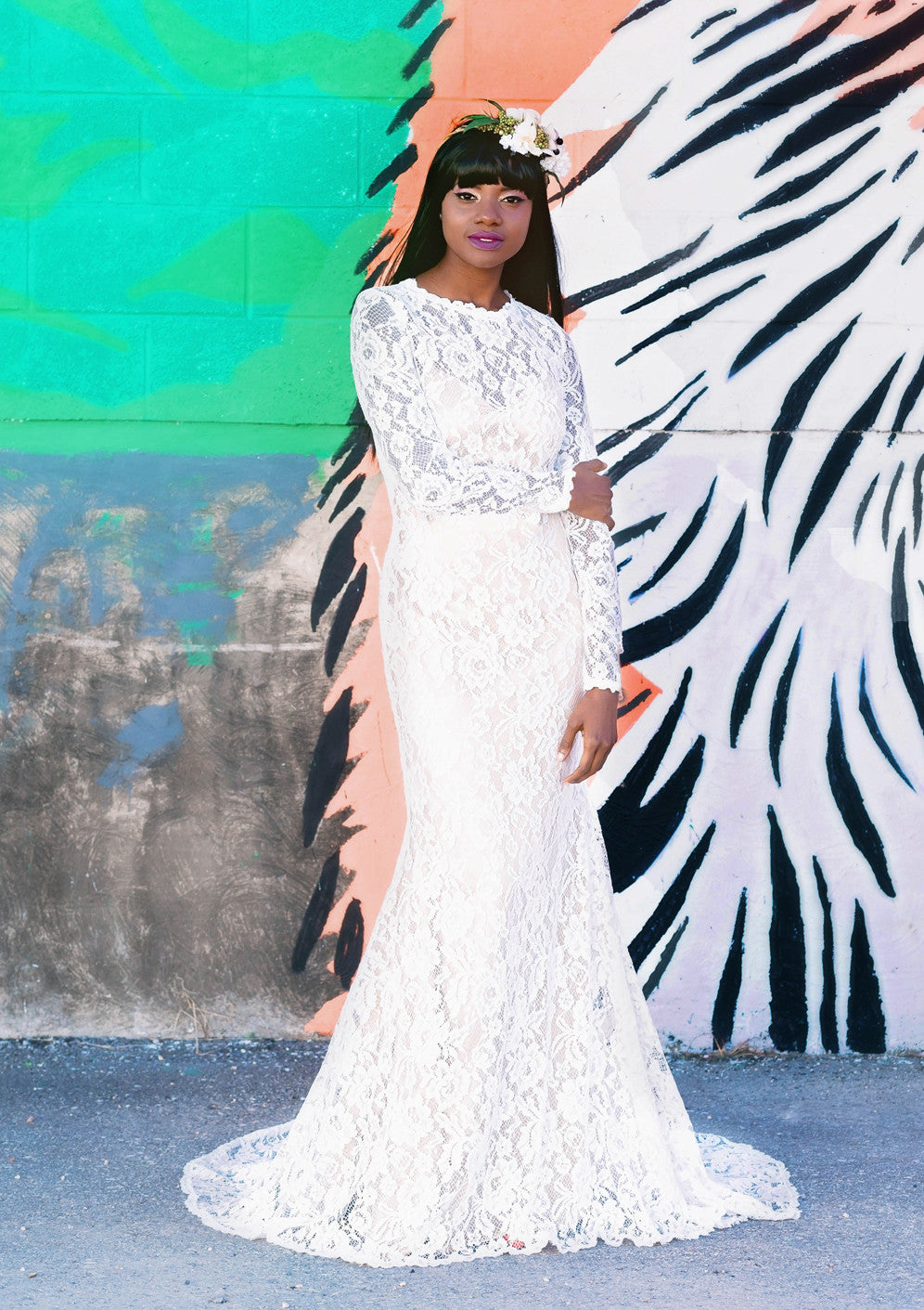 Bride in white lace mermaid wedding dress with long sleeves, floral crown, against colorful mural wall
