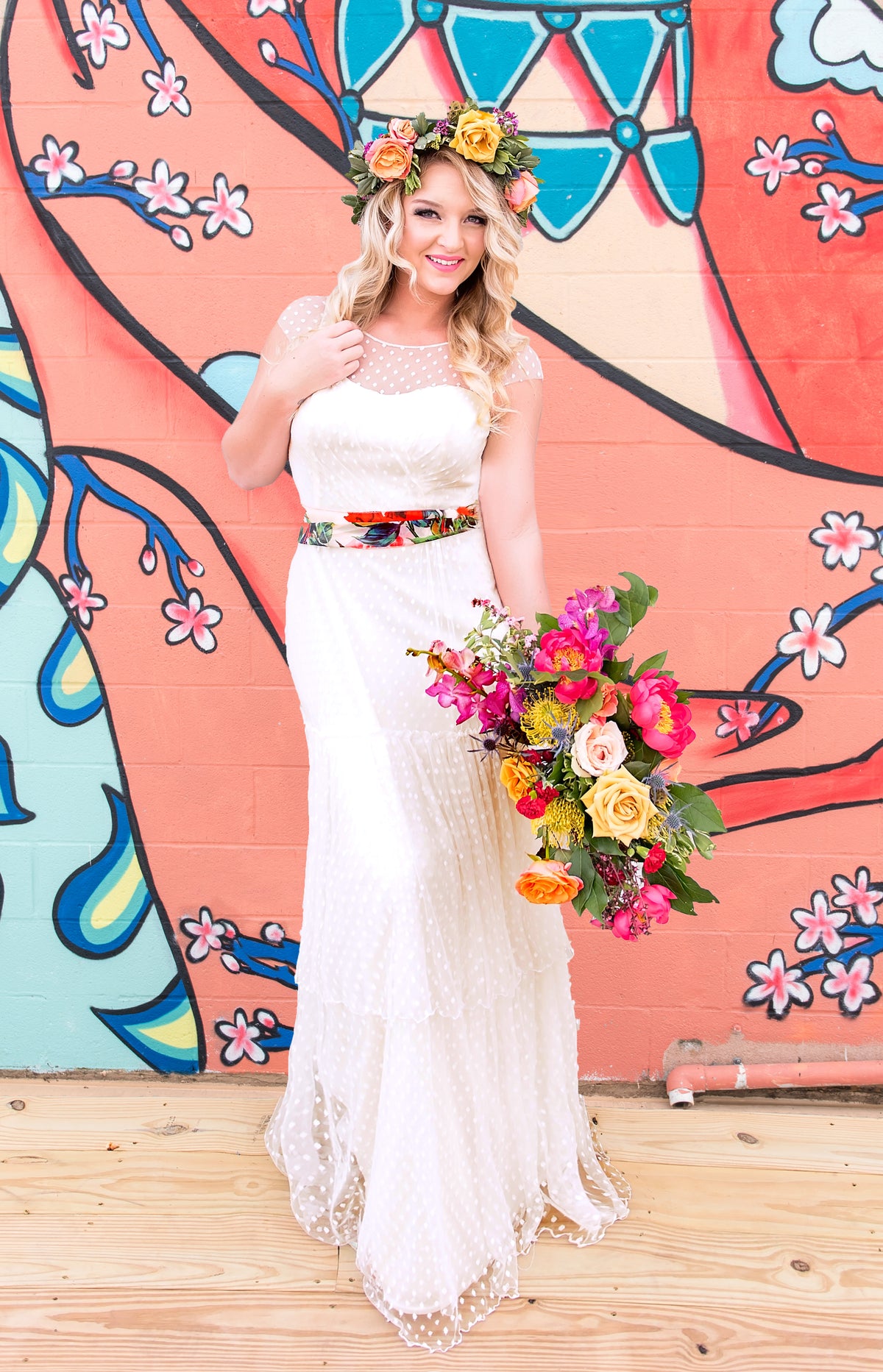 Bride in polka dot wedding dress with floral crown and bouquet, standing before colorful mural
