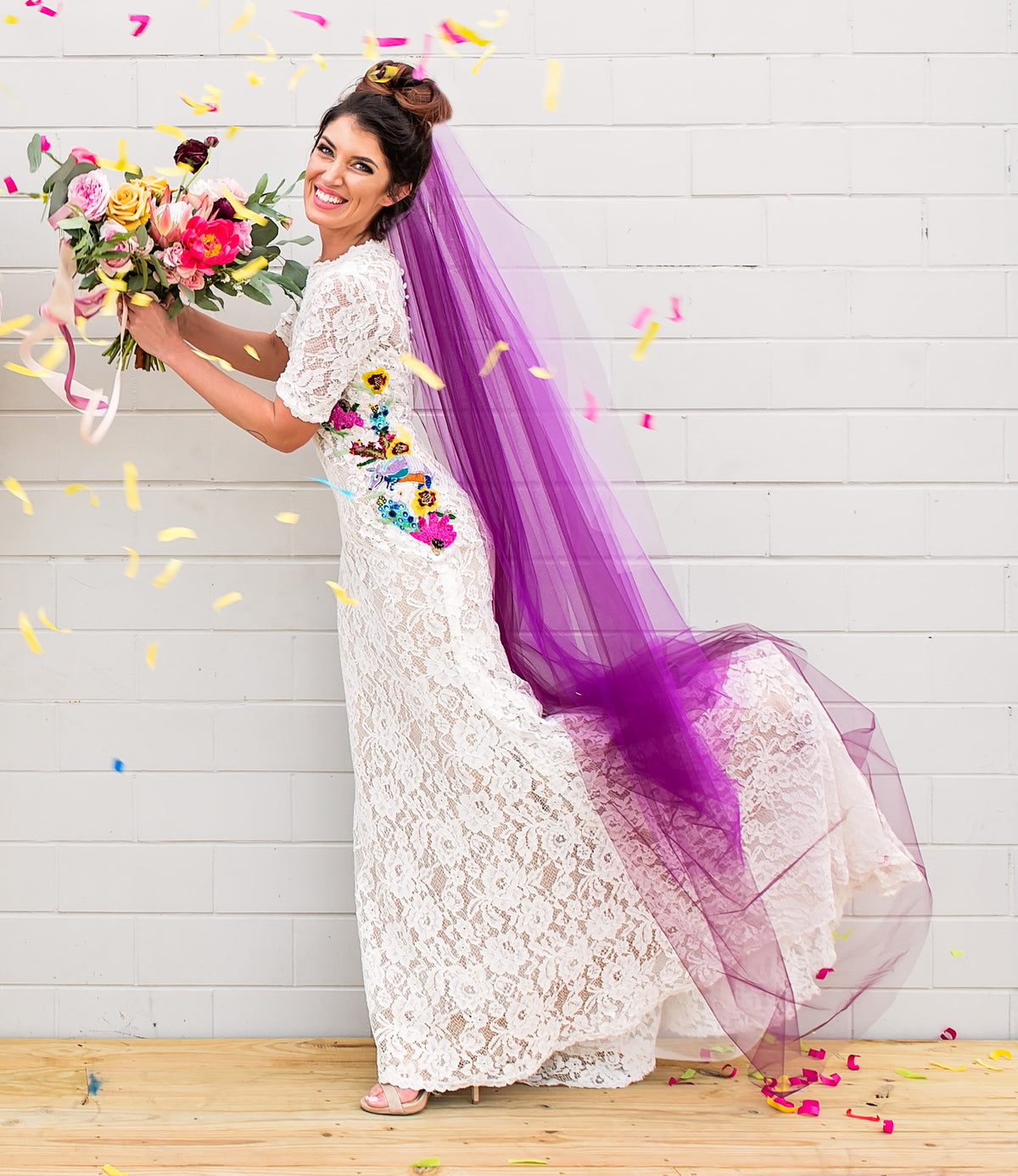 Bride in white lace wedding dress with colorful floral embroidery, purple veil, and confetti, holding bouquet
