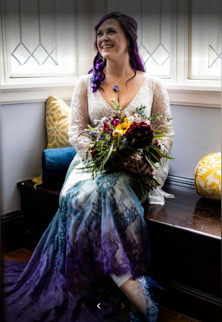 Bride in ombre lace wedding dress with purple hair, holding colorful bouquet, seated by window