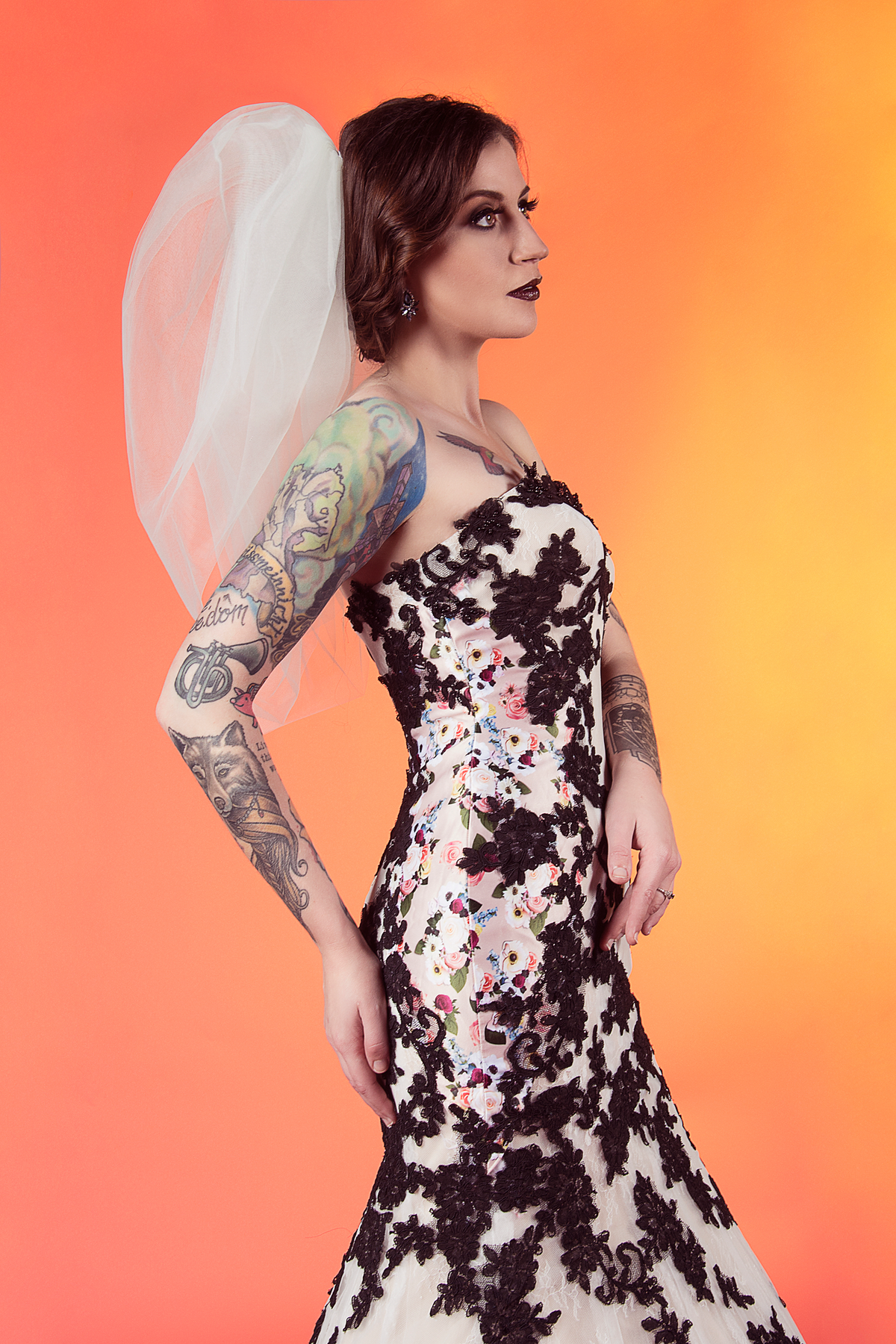 Tattooed bride in floral lace wedding dress with veil, bold makeup, and vintage style against warm gradient background