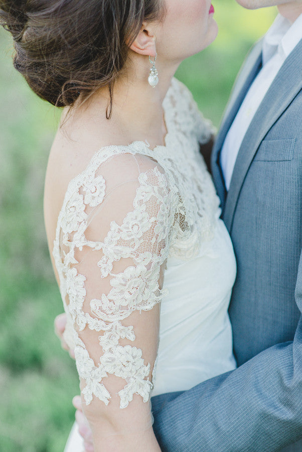Bride in lace wedding dress with floral detailing, embracing groom in gray suit outdoors