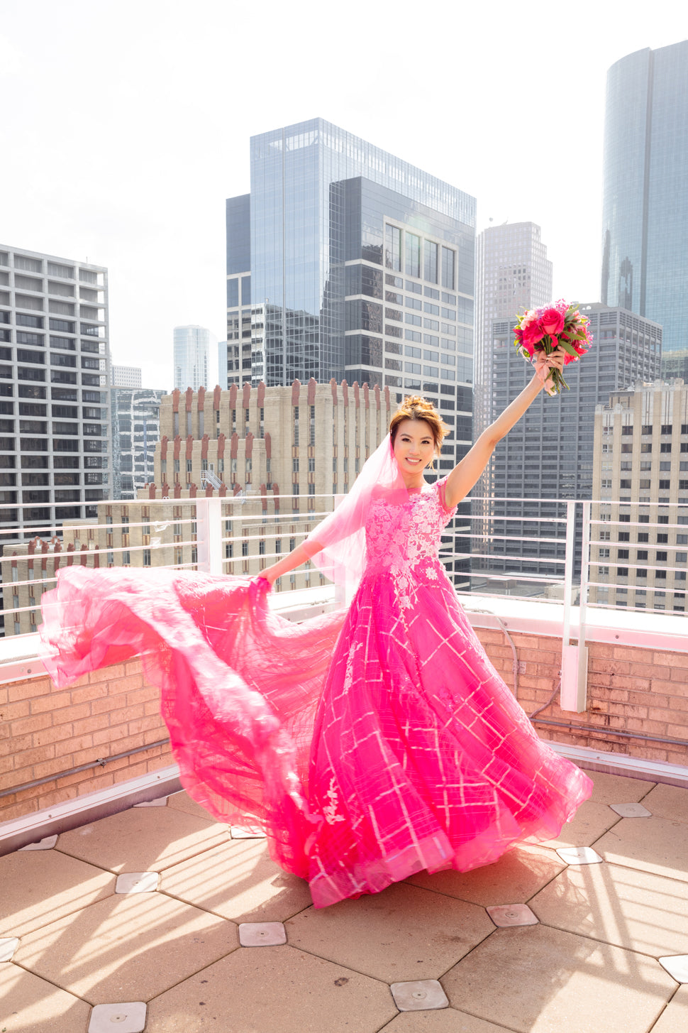 Bride in vibrant pink custom wedding dress with flowing skirt, holding bouquet on city rooftop