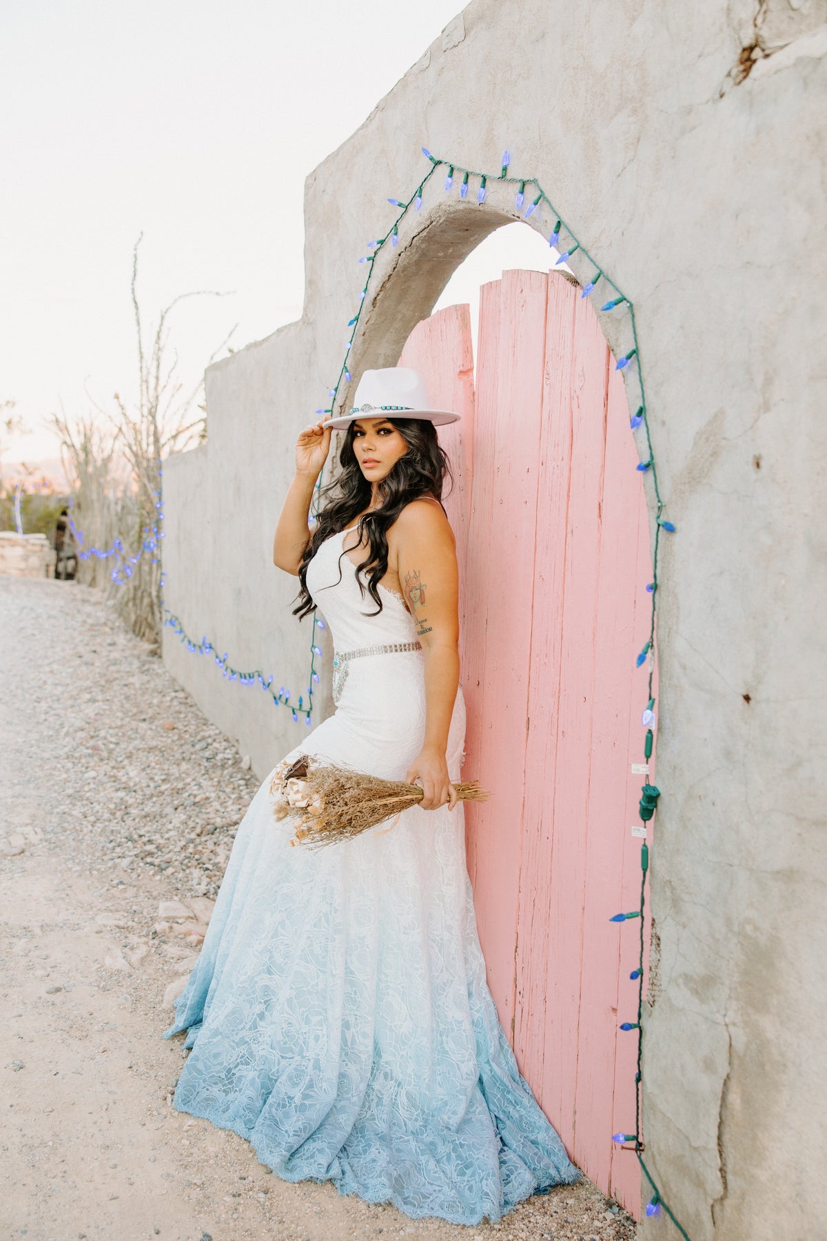 Bride in ombre lace wedding dress with white hat, holding dried bouquet by pink door with blue lights
