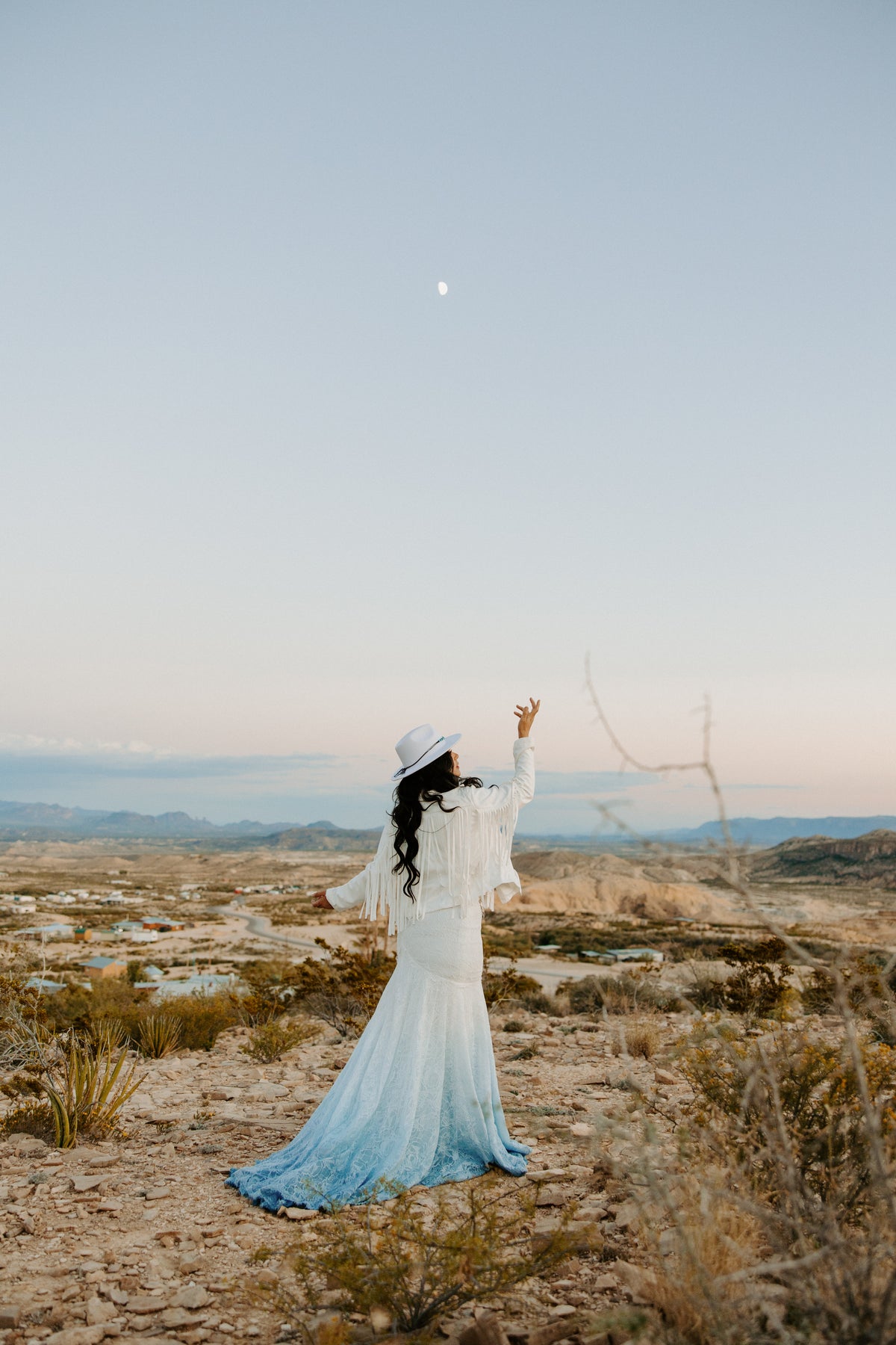 Bride in ombre blue lace wedding dress with fringe jacket and white hat, desert sunset