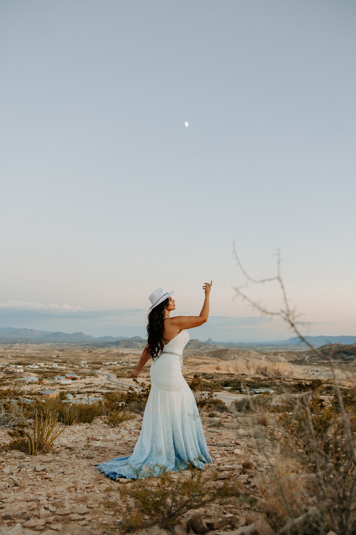 Bride in ombre blue wedding dress & white hat, reaching toward moon in desert landscape
