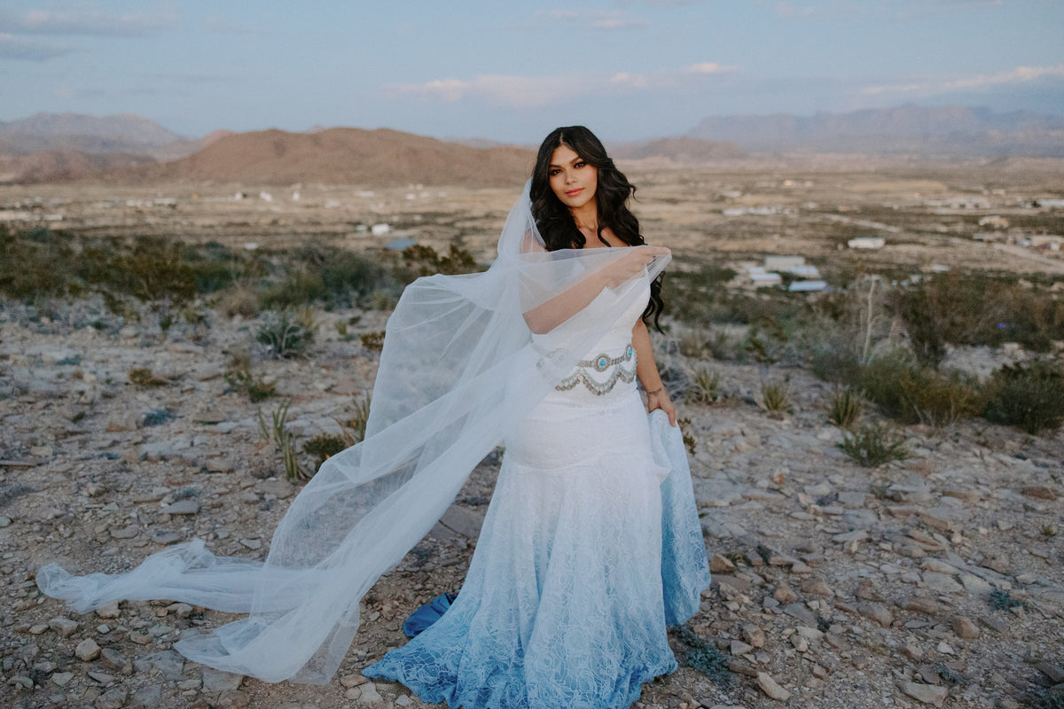 Bride in custom ombre blue lace wedding dress with flowing veil, desert landscape backdrop