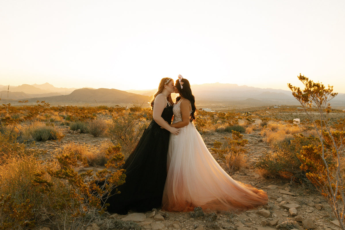 Two brides kiss in desert sunset, one in black wedding dress, other in ombre pink-to-white gown, mountain backdrop