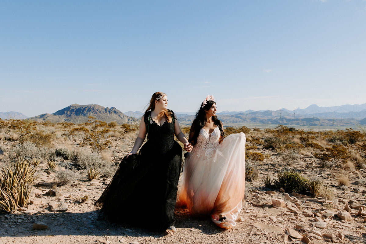 Two brides in custom wedding dresses, one black and one ombre pink, holding hands in a desert landscape with mountains.